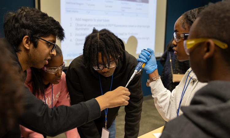 A presenter teaches a student to operate a pipette with three other students standing in a circle watch and listen.